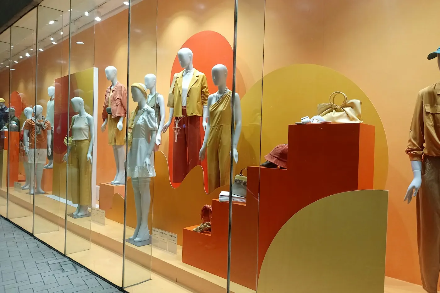 Shop window display with mannequins on orange and yellow stepped plinths, showing retail entrance and display branding